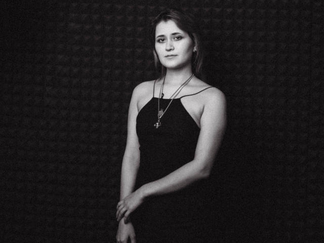 Photo en noir et blanc d'AlexaLexie, avec une poitrine de taille moyenne, portant une robe noire, un collier avec une croix, les bras croisés, un fond sombre et texturé, une expression légèrement agacée, des cheveux bruns de longueur moyenne..