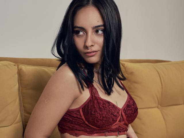 AshleyDaphna with black hair, nose ring, and red lace bra, showing her small tits, sitting on a yellow couch. Her arms are relaxed, and she's giving a subtle smile.