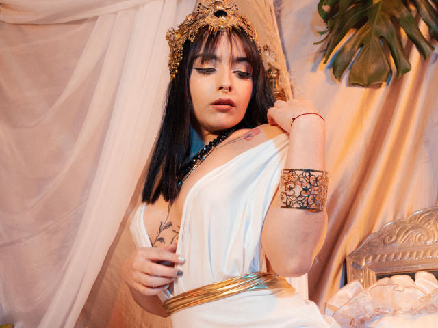 AshleyDaphna with black hair, gold headpiece, white dress, gold belt, arm bracelet, tattoo, and nose ring, looking down, holding dress, soft pink and gold background.