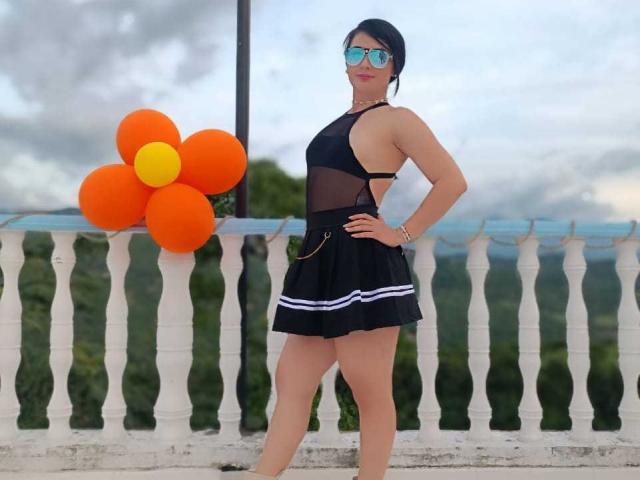 FrancheskaWaalsh standing with hands on hips, wearing black sheer top, black skirt with white stripes, blue reflective sunglasses, black hair in bun, orange and yellow balloon to left.