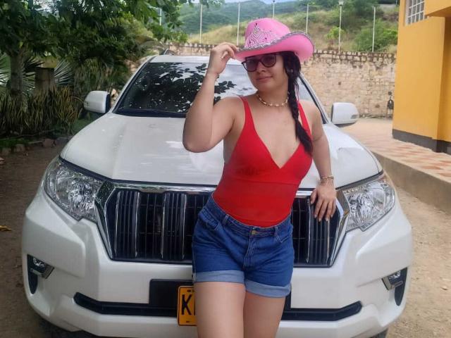 FrancheskaWaalsh with pink cowboy hat, red tank top, showing her boobs, blue denim shorts, standing in front of white Nissan car, wearing glasses, necklace, and bracelet.