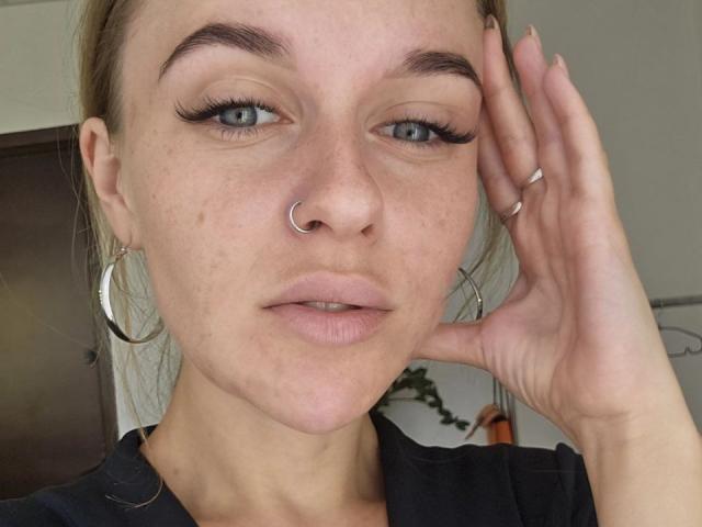 MaribelRiverr has light freckles, blue eyes, and thick eyelashes. She's wearing a nose ring, hoop earrings, and a black shirt. Her hand is touching her head.
