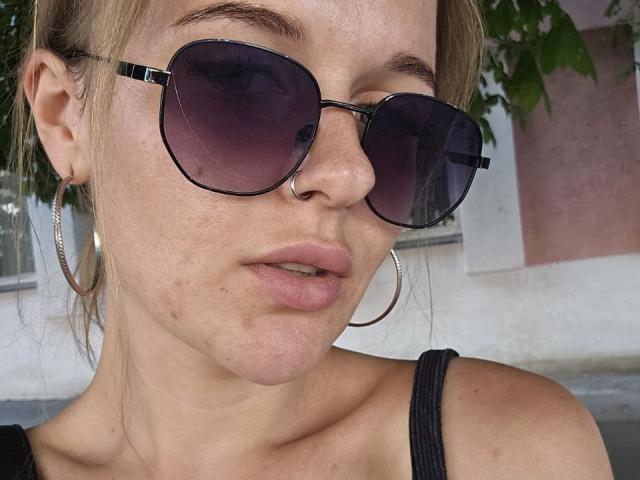 MaribelRiverr has a nose ring, wearing large silver hoop earrings and dark purple aviator sunglasses, with a black tank top, showing her pinkish lips and slightly freckled face.