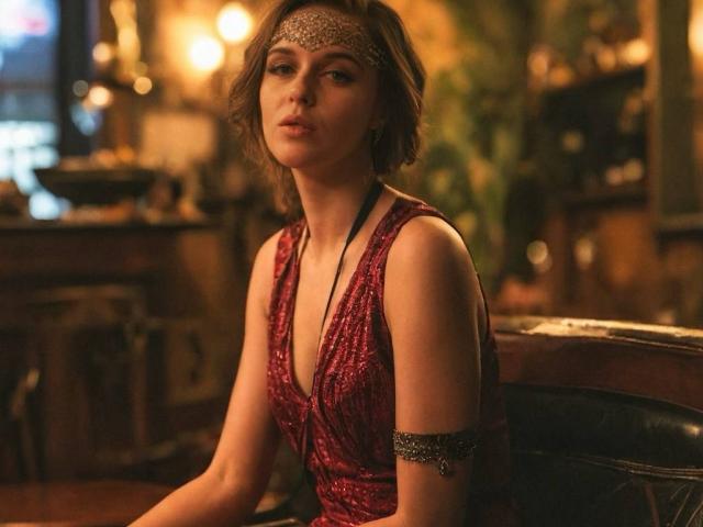MaribelRiverr, wearing a red, sparkly dress with a deep V-neck, silver headpiece, and arm bracelet, sits in a dimly lit room with a sexy, confident look.