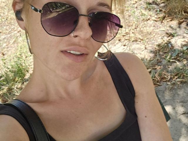 MaribelRiverr with round sunglasses, black tank top, nose ring, and earbuds, showing a slight smile, sunlight on her face, and a black strap over her shoulder.