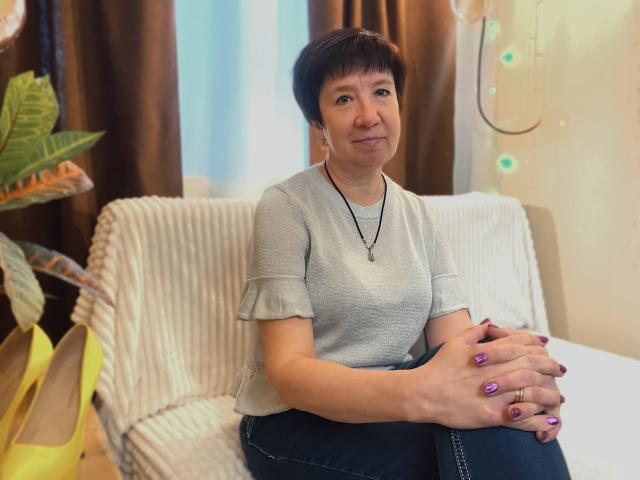 EllaPork, short brown hair, gray ruffled shirt, black jeans, purple nails, hands on lap, necklace, sitting on white striped couch, yellow bag, plant, smiling, cozy indoor setting.