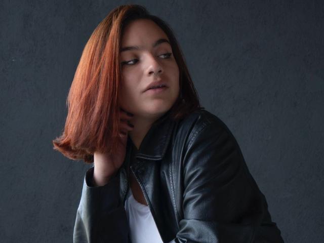 SabriinaMorgan with short, reddish-brown hair, wearing a black leather jacket over a white top, looking to the side with a slightly annoyed expression. She's touching her neck.