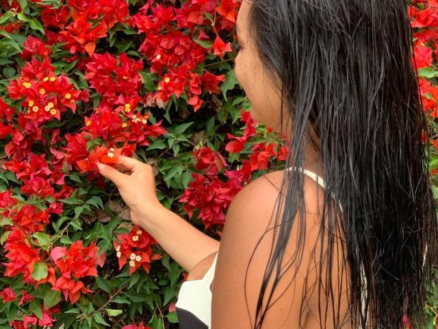 Gala Malkova mit nassen, schwarzen Haaren, die leuchtend rote Blumen mit gelben Blüten berührt, trägt ein weißes und schwarzes Oberteil, vor einem üppigen Hintergrund aus grünem und rotem Laub..