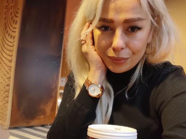 Katrina, avec ses cheveux blonds, son col roulé noir et sa montre dorée, souriant tout en tenant une tasse de café, une main sur sa joue, portant des boucles d'oreilles pendantes, avec des yeux marron et de longs cils..