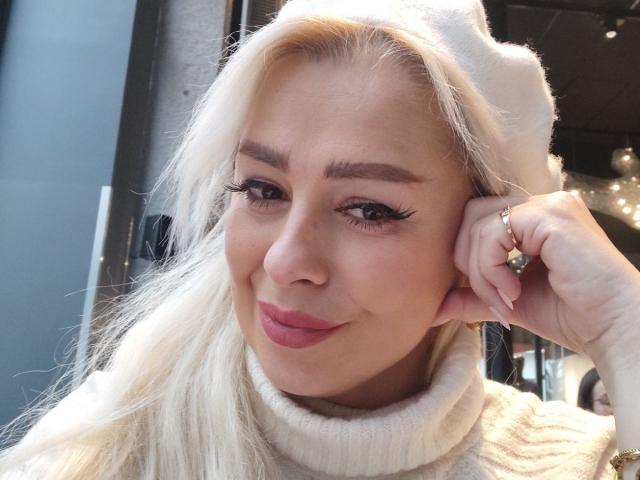 KatrinaCharm with blonde hair, white hat, and pink lipstick, smiling slightly, wearing a white turtleneck, holding her ear with a ring, showing long eyelashes.