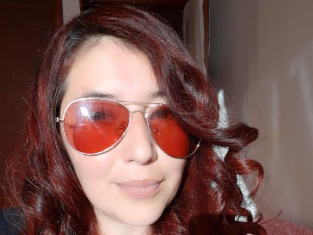 AntonnellaS, avec ses cheveux roux bouclés, porte des lunettes de soleil aviateur teintées de rouge. Elle a une peau claire et un léger sourire, et son petit nez et ses lèvres sont visibles..