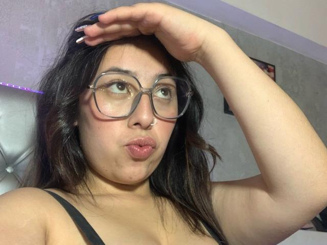 ScarlettParchMusa with glasses, pouty lips, black hair, fair skin, black tank top, hand in hair, looking up, purple light, white wall, framed picture, purple strip light.
