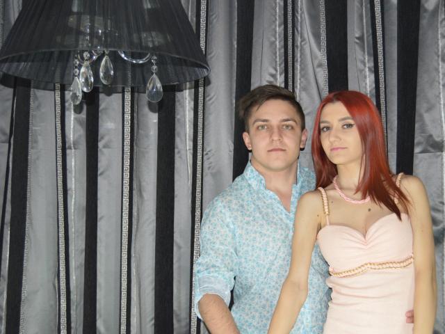 Pareja atractiva con cabello rojo, pechos pequeños, vestida de rosa, de pie junto a otra pareja atractiva con cabello castaño, camisa con estampado azul, que parece seria. Una lámpara negra con gotas de agua en el fondo..