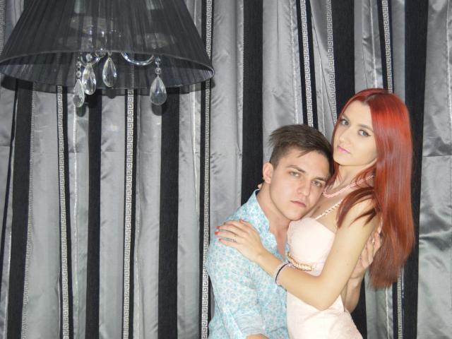 HotCoupleAnal with red hair, tits in pink bra, stands next to HotCoupleAnal with brown hair, small ass, in blue shirt, arms around each other, under black lamp.