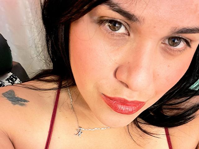 VivianaThompson has red lipstick, brown eyes, black hair, and a butterfly tattoo on her left shoulder. She's wearing a red spaghetti strap top and a silver necklace.