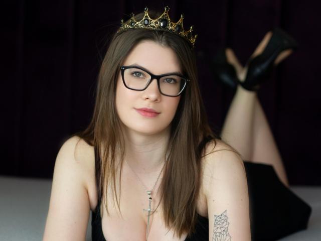 CarolineRose with glasses, brown hair, black crown, black dress, necklace, cross, tattoo on right arm, black heels, legs up, smiling, pale skin, pink lipstick.