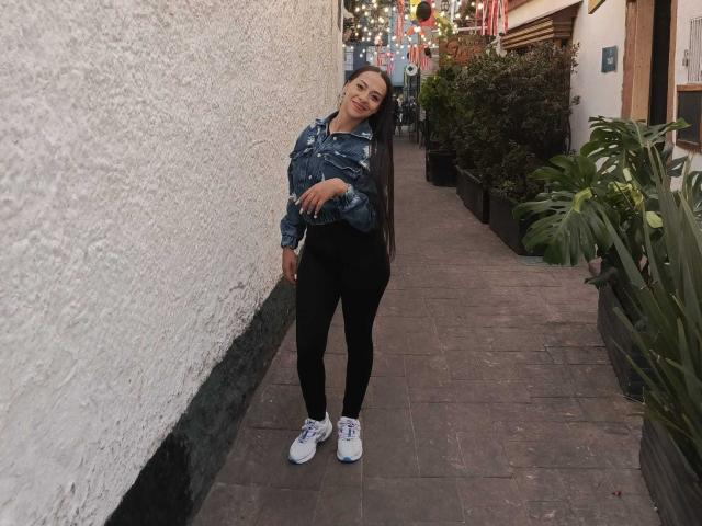 MyaLoreen stands leaning against a white wall in a narrow alley. She wears a denim jacket, black pants, and white sneakers, smiling with her arm across her chest.