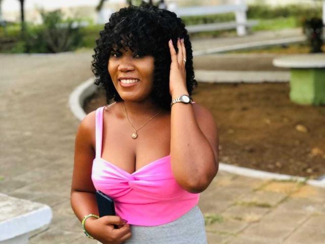 Davlov with curly black hair, wearing a pink top showing her tits and a gray skirt, smiling, hand in her hair, pink top, gold necklace, watch, green bracelet.
