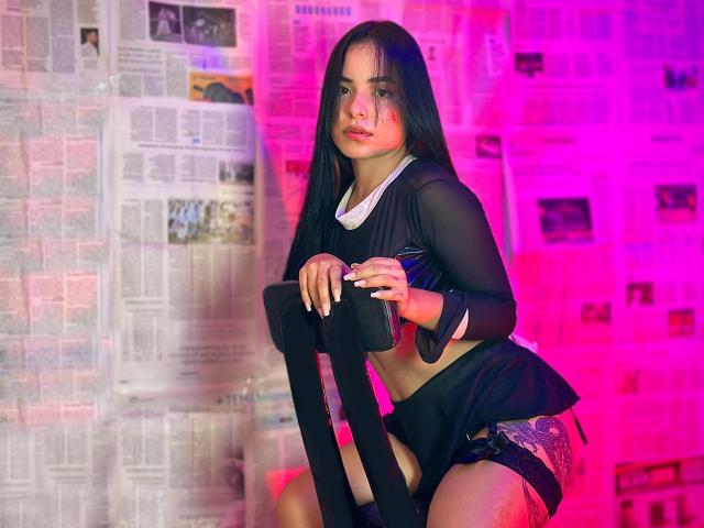 ScarletFoxxi, com longos cabelos pretos, vestindo uma blusa preta transparente que revela seus pequenos seios, shorts pretos e meias até a coxa, agachada em uma cadeira, sob iluminação rosa e roxa..