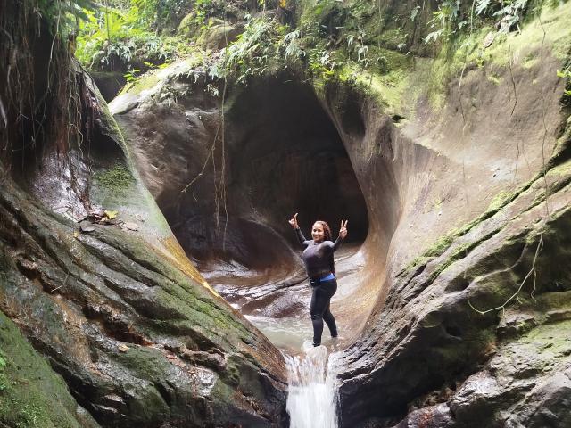 YiyaHot69 is jumping through a rocky cave with a small waterfall, arms raised, wearing a dark shirt and black pants, surrounded by green moss and hanging plants.