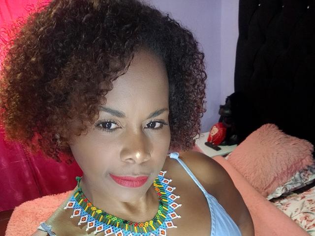Afrodita Kane, con i capelli castano ricci, indossa una collana colorata fatta di perline e un top blu a spalline sottili. Ha il rossetto rosso e un leggero sorriso, che mette in mostra la sua pelle marrone media e il seno..