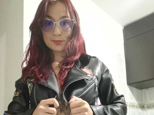Bellanid with red hair, wearing a black leather jacket, holding a dark brown, erect penis, and smiling with red lipstick, wearing purple-tinted sunglasses.