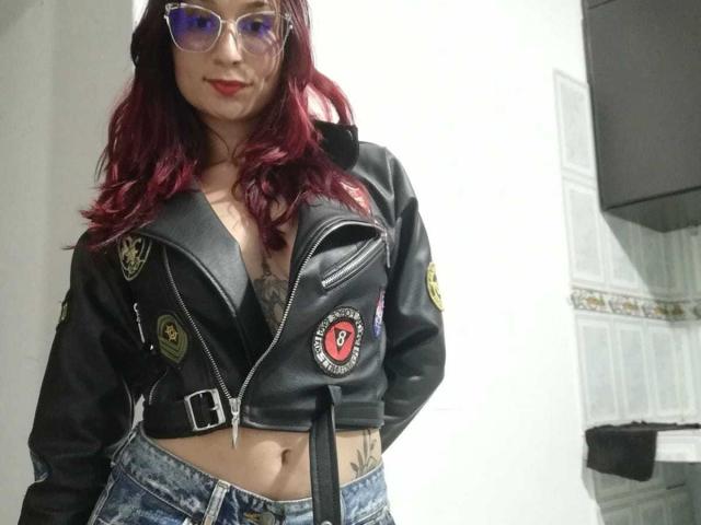 Bellanid with red hair, glasses, black leather jacket, exposing her tits, tattoo on her side, wearing blue jeans, standing with hands behind back.