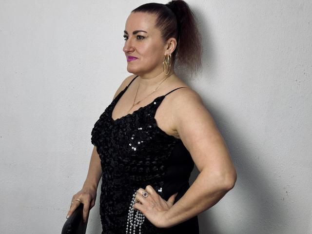 DianaMGG tiene un trasero regordete, lleva un vestido negro con lentejuelas, aretes de aro dorados y una coleta. Está de pie con una mano en la cadera, sosteniendo un bolso negro..