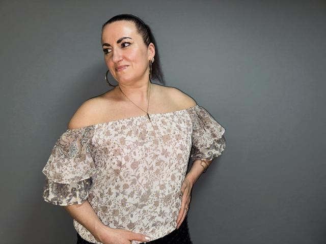 DianaMGG with a lace off-shoulder top, showing her tits slightly, dark hair in a ponytail, hoop earrings, hands on hips, smiling, gray wall in background.