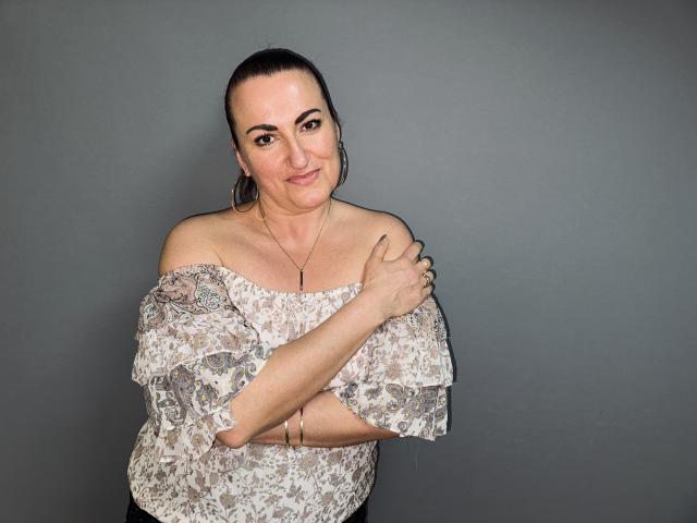 DianaMGG with dark hair in a ponytail, wearing an off-shoulder floral top, arms crossed over her chest, small tits, subtle smile, gray background, earrings, and a necklace.