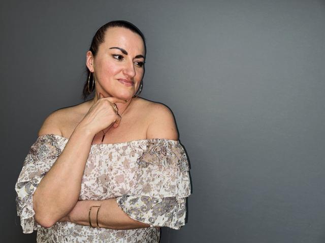 DianaMGG with a slight smirk, brown hair in a ponytail, off-shoulder patterned top, gold hoop earrings, arm bracelets, hand touching chin, gray background.