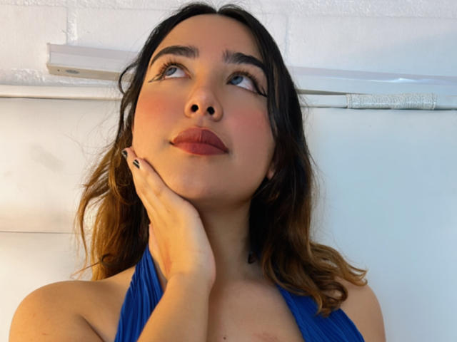 SwetiieBoobs com cabelo castanho ondulado, top azul de alças, mão no queixo, olhando para cima com lábios rosados, olhos azuis e maquiagem discreta, em frente a uma parede branca..