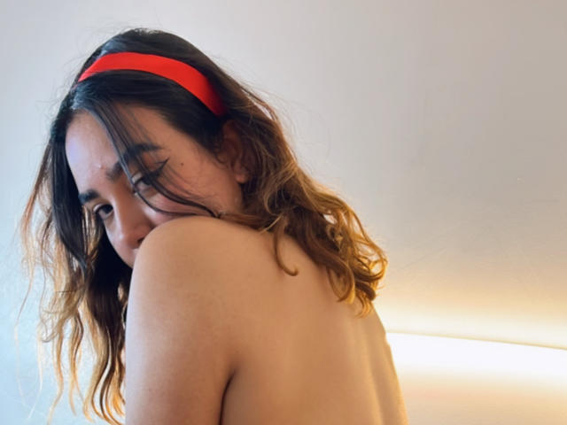 Topless, SwetiieBoobs met golvend bruin haar, een rode hoofdband en een verlegen glimlach, die haar blote schouders laat zien en over haar schouder kijkt. Zacht licht van onderen..