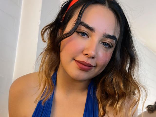 SwetiieBoobs tem cabelos castanhos ondulados com pontas loiras, veste um top azul de alças e tem uma expressão adorável, ligeiramente convencida, com batom vermelho e delineador de olhos..