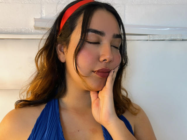 SwetiieBoobs com os olhos fechados, uma faixa vermelha na cabeça, um top de alça azul, a mão no rosto, cabelo castanho ondulado, blush rosa suave, um leve sorriso, uma parede de tijolos brancos ao fundo, uma atmosfera relaxante..