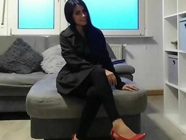 Amelia Miranda, con cabello negro largo, vestida con un abrigo negro, pantalones negros y zapatos planos rojos, sentada en un sofá gris, con las piernas cruzadas, sonriendo, en una habitación con paredes blancas..