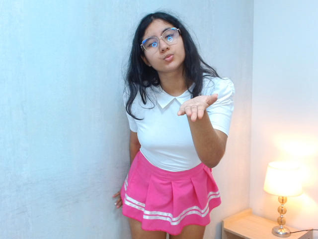 TereCarpert, con cabello largo y negro, gafas azules, camisa blanca, falda rosa con rayas blancas, señalando, haciendo pucheros, de pie en una habitación con una lámpara sobre una mesa de madera..