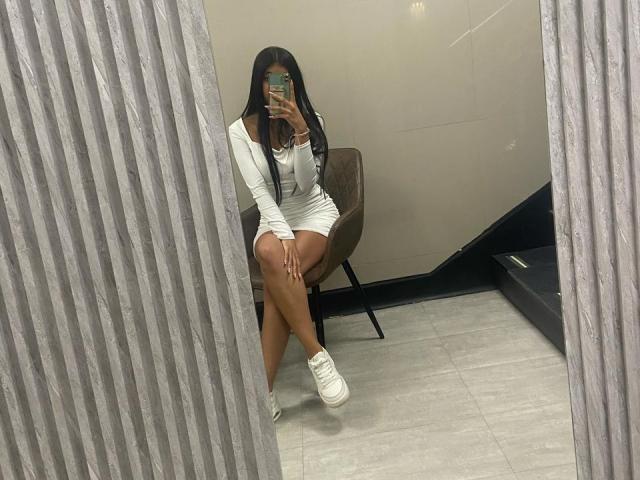 EmilyBanner, con lunghi capelli neri, vestito bianco aderente che mette in mostra il sedere, seduta, che tiene un telefono, scarpe da ginnastica bianche, in un selfie allo specchio, gambe incrociate, parete grigia a coste, sedia marrone..