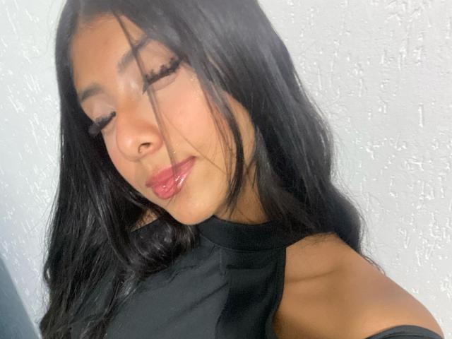 EmilyBanner, con lunghi capelli neri, occhi chiusi, labbra rosa lucide, indossa un top nero con spalline, ha la pelle chiara e un trucco leggero, sullo sfondo di una parete bianca con una texture..