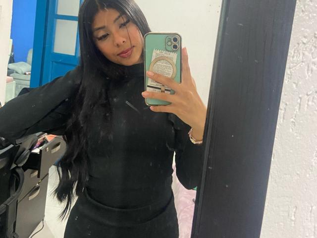 EmilyBanner, con cabello negro largo, vestida con un cuello alto negro ajustado, tomando una selfie en el espejo, mostrando sus senos, sosteniendo un teléfono verde, mirando a la cámara, con lápiz labial rojo..