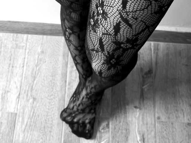 MaddieHottie's black lace-patterned legs standing on a wooden floor, with her feet slightly apart, showing her bare toes and the intricate design of the lace stockings.