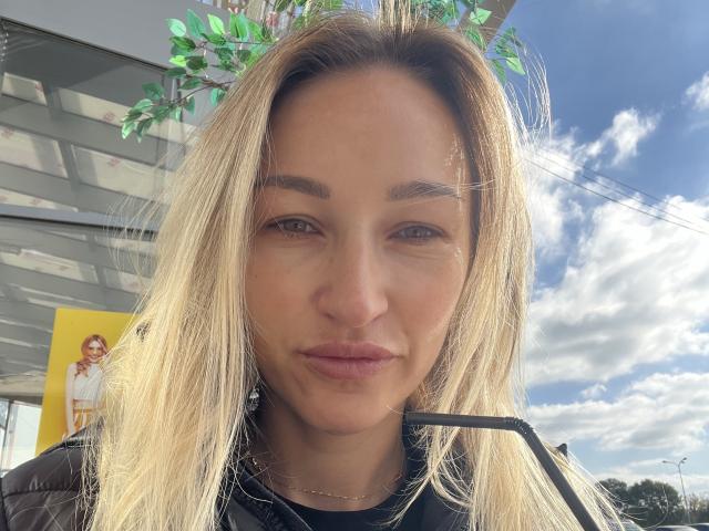 Lilitraya has blonde hair, brown eyes, and a serious expression with slightly puffed lips. She's wearing a black puffy jacket and has a leafy branch in her hair.
