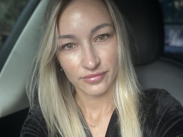 Lilitraya with blonde hair, green eyes, and pink lips, wearing a black top, has a subtle smile, and a thin gold necklace, sitting in a car.