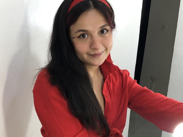 Miilyhot with long black hair, red headband, and red shirt, smiling, has a small heart-shaped makeup mark under her right eye, and a cute, petite face.