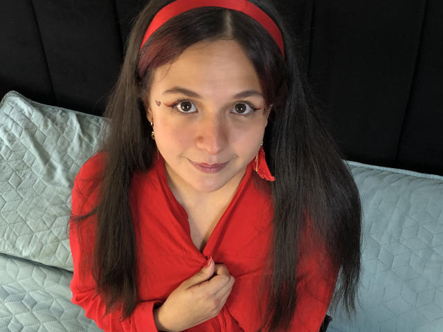 Miilyhot with long dark hair, red headband, red shirt, small tits, holding shirt, cute face, brown eyes, red earrings, sitting on light blue quilted bed, black headboard.