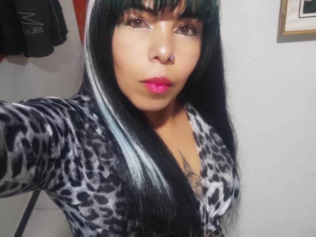 AvelynNoir with long black and white hair, nose ring, pink lipstick, leopard print robe, tattoo visible below chest, taking a selfie. Boobs slightly visible under robe.