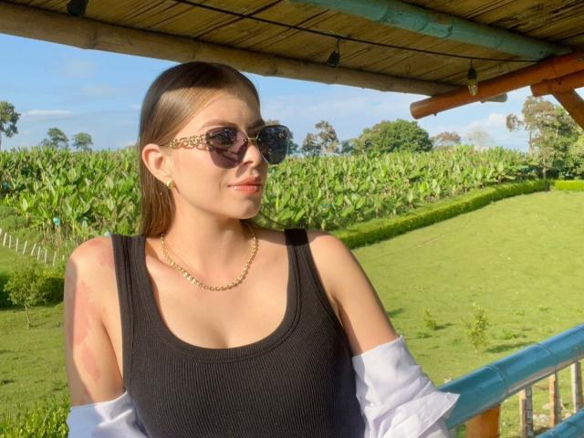 AnnaFer mit braunen Haaren, die eine Sonnenbrille, ein schwarzes Tanktop und eine goldene Kette trägt, steht draußen. Ein weißes Hemd ist von ihren Schultern gerutscht. Im Hintergrund ist ein grünes Feld und Bananenpflanzen zu sehen..