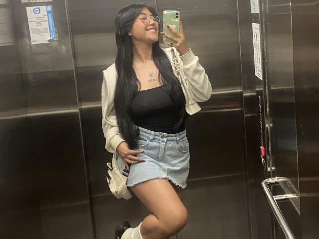 Elizaa met lang zwart haar, een bril, een witte cardigan, een zwarte top en een spijkerrok, maakt een spiegelzelfportret in een lift, waarbij haar borsten en billen zichtbaar zijn, en ze glimlacht..