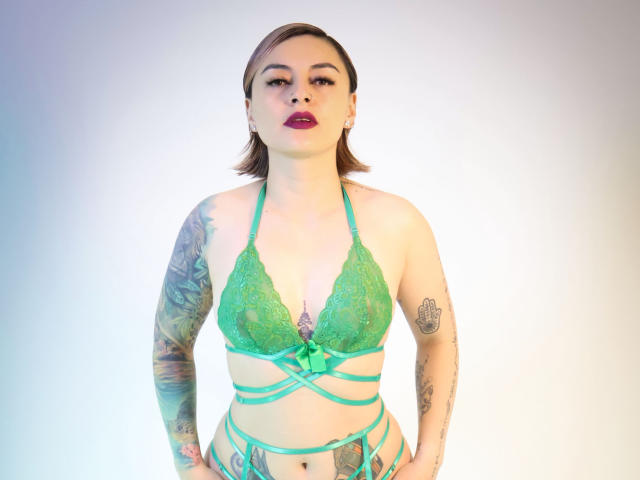 KatParadise with tattoos, green lace bra, and strappy green lingerie, standing confidently, hands on hips, red lipstick, brown hair, looking directly at camera.