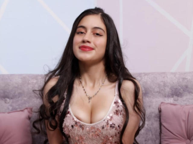 AnyeliHoot, com cabelos pretos longos, batom vermelho e um vestido rosa com lantejoulas que mostra seus seios grandes, sorrindo, usando um colar, sentada em um sofá rosa e cinza..